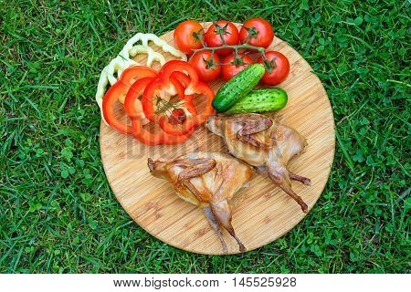 Delicious fried quail with fresh juicy vegetables on the wooden round board on green grass. Prepared tomato, cucumber, sweet pepper, partridge, quail. 