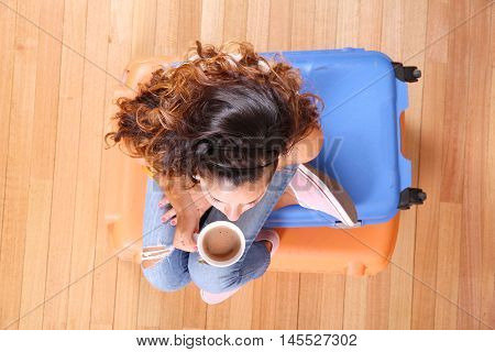 A young woman sitting on a stack of suitcases while drinking coffee and waiting for the departure to vacations.