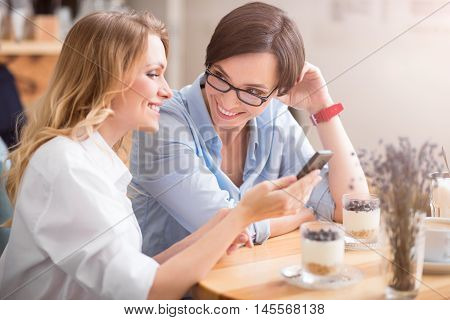 Friendly ladies. Positive and happy women holding smart phone while sitting at table in cafeteria and telling secrets