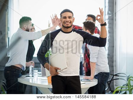 Picture of many business colleages or office workers showing victory of teamwork on background while handsome businessman posing with his hand in pocket in office interior.