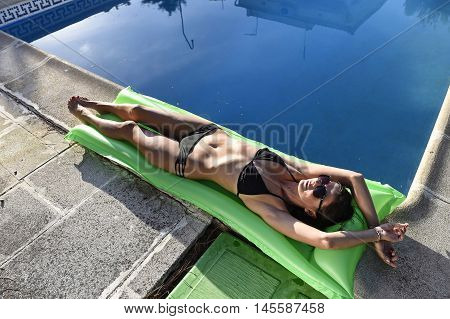 young sexy woman in bikini and sunglasses with fit beautiful body having suntan relaxing on airbed at swimming pool of vacation hotel resort in summer enjoying holidays