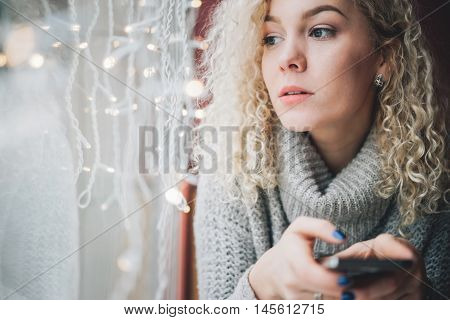 Young blond curly female in warm sweater seating in cafe, thoughtfully looks into the distance, holding a smartphone, winter city outside the window