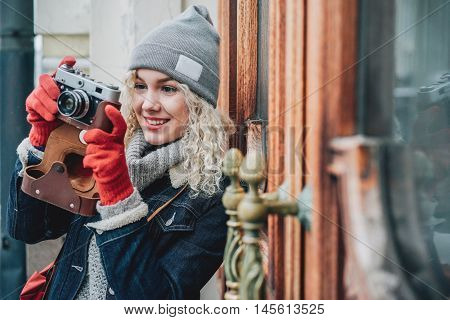 Young blond curly female in warm clothes with old film camera shooting a photo on the background of wooden doors with handles, winter, snow
