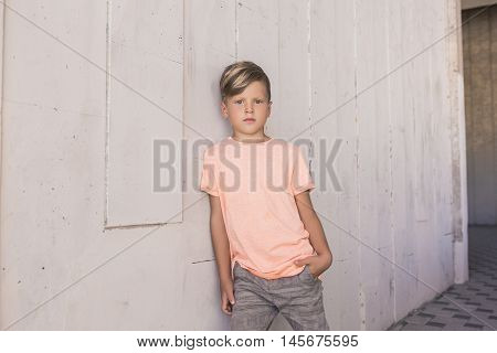 Trendy boy leaned on the wall and is confidently looking forward . He has cool hair . With one hand he put in his pocket . Children's fashion