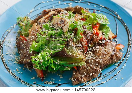 Restaurant food closeup. Warm meat salad with sesame, vegetables and lettuce on blue plate. Appetizing dish served with soy sauce, dinner meal. Decorated healthy meal for gourmet