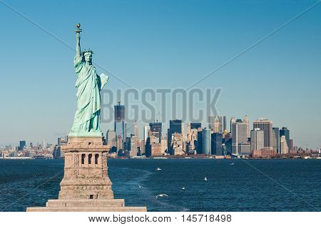A vantage point in New York Harbor the iconic island of Manhattan lies just ahead. An eclectic mix of buildings spreads across the city creating a colorful skyline. In the foreground just ahead and to the left the Statue stands guard.