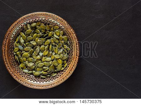Raw pumpkin seeds in an antique meral bowl on a dark textured background