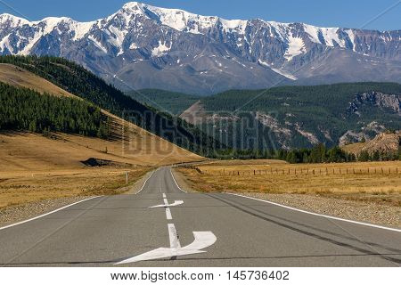 Scenic view with the asphalt road in the steppe going to the mountains with snowy peaks and slopes covered with forest on a sunny day