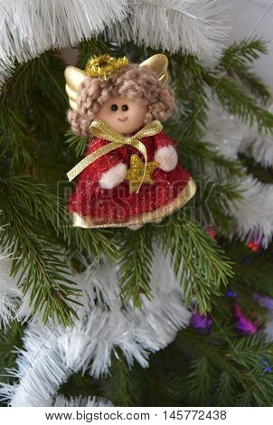 Christmas toy in the form of an angel. The toy is located on a fir-tree. An angel in red clothes. The angel holds a star