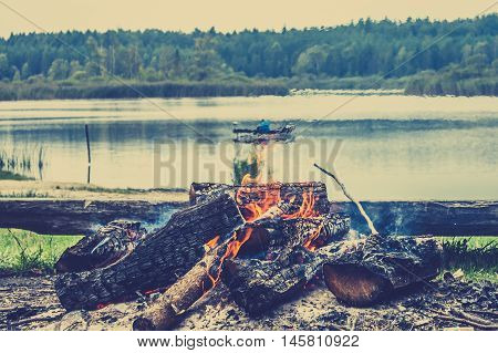 Rustic bonfire by the lake in the forest on the background of blurry boat with fisherman on the lake vintage photo.