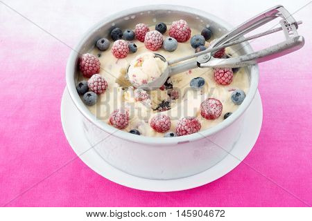 Homemade cheesecake ice cream with mixed berries in a round metal form decorated with blueberries and raspberries and with an ice cream scoop in the middle. Pink clothes as background.