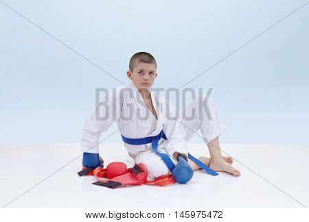 On a light background karate athlete sits near a karate outfit