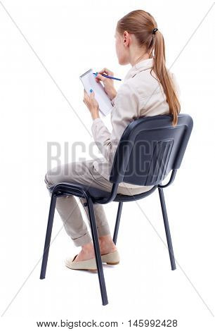back view of young beautiful woman sitting on chair and takes notes in a notebook. Isolated over white background. Skinny girl in white denim suit thoughtfully sidit on a chair taking notes in a