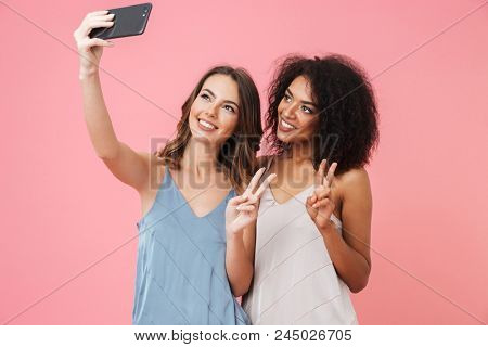 Two pretty young girl in dresses taking a selfie while showing peace geture isolated over pink background