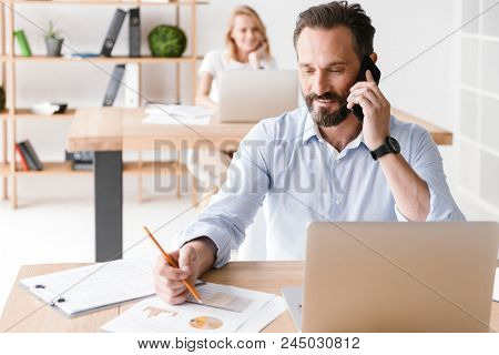 Smiling man manager working with laptop computer and documents while sitting at the office and talking on mobile phone with woman co worker on a background