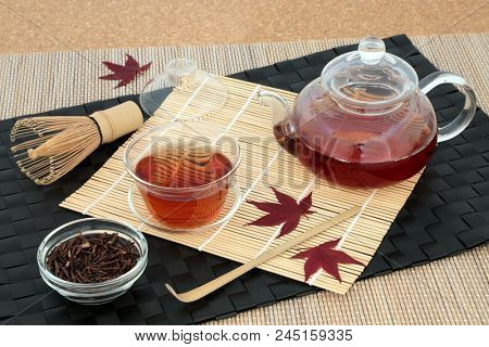 Japanese kuchika roasted twig green tea with glass teapot & cup, dried twigs in a bowl, stirrer, whisk & maple leaves on bamboo background. Has many health benefits including rejuvenating properties.