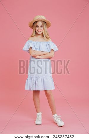 Full length portrait of beautiful blonde summer girl 8-10 wearing blue dress and straw hat posing at camera with arms crossed and smile isolated over pink background
