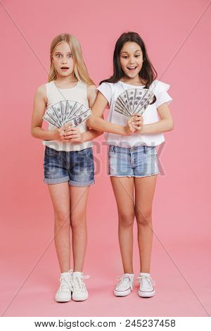 Full length image of delighted brunette and blonde girls 8-10 wearing casual clothing expressing surprise while holding lots of money in dollar cash isolated over pink background