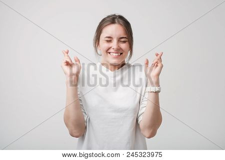 Indoor Shot Of Attractive European Girl In White Shirt Having Excited Look, Keeping Fingers Crossed,
