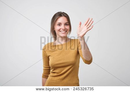 Friendly-looking European Teenager Dressed In Yellow Pulover Saying Hello, Waving Her Hand. Positive