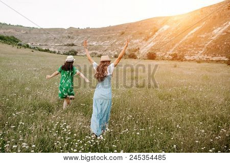 Two Attractive Happy Young Woman Walking On Field With Flowers Through The Mountain On The Sunset. F