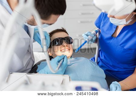 medicine, dentistry and healthcare concept - dentist and assistant with dental drill and saliva ejector treating kid patient teeth at dental clinic
