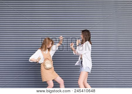 Two Young Girlfriends In Stylish Clothes Dance On A Gray Background. Portrait Of Two Cheerful Girls 