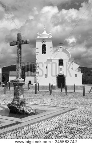 Black And White Image Of The Cross And Church In The Main Square Of Querenca, Portugal