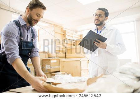 Handsome Bearded Warehouse Worker Packing Detail While His Superior Wearing Lab Coat Keeping Eye On 