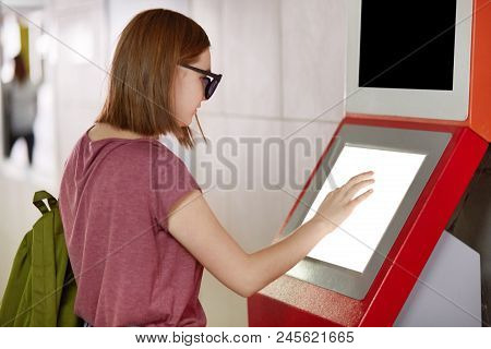 Sideways Shot Of Pretty Female Carries Rucksack, Wears Shades, Stands In Front Of Terminal, Tries To