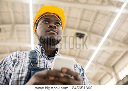 Below View Of Serious Introspective Young African Construction Engineer In Yellow Hardhat Texting Me