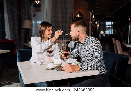 Young Couple Feeding Each Other In Restaurant, Handsome Man And Beautiful Woman Are Sitting At The T