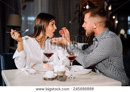 Young Couple Feeding Each Other In Restaurant, Handsome Man And Beautiful Woman Are Sitting At The T
