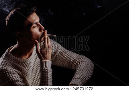 Thougtful Young Man, Wearing Warm Knitted White Sweater Smoking Sigarette, Thinking On Dark Backgrou
