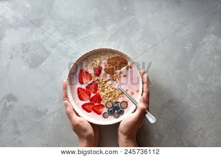 Plate with a spoon and an apetitic yoghurt with berries and almonds women's hands holding on a gray concrete background