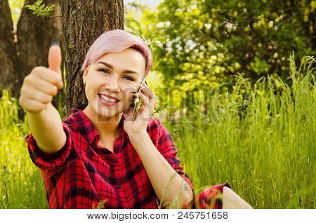 Young Beautiful Smiling Girl With Pink Hair Sitting In Green Grass, Call By Smartphone And Shows Thu