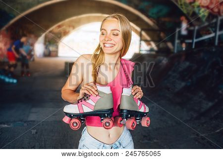 Blonde Girl Is Smiling. She Is Standing In A Training Room For Rollerbladers. Rollers Are Hanging Ov