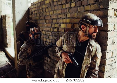 A Picture Of Serious Bearded Man Hiding Behind Wall And Looking To The Side. He Has Gun. Other Two M