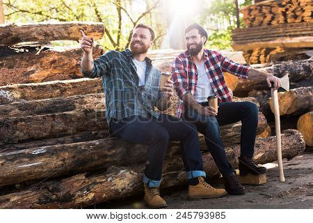 Smiling Lumberjack With Coffee Cup Pointing By Finger To Partner With Axe Sitting On Logs At Sawmill