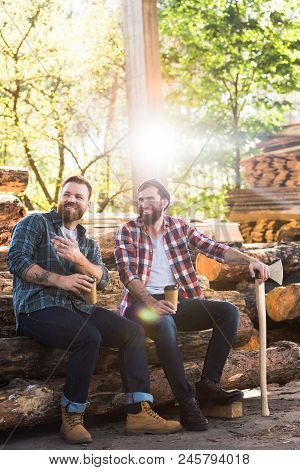 Lumberjack With Coffee Cup Pointing By Finger To Partner With Axe Sitting On Logs At Sawmill