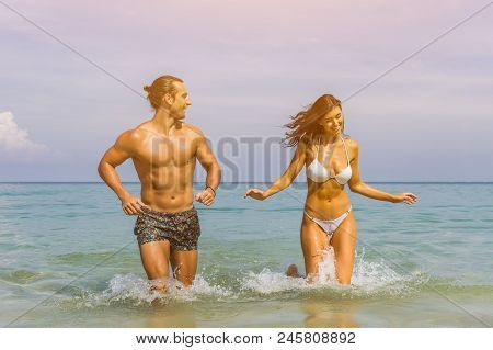 Happy Couple Laughing Together Holding Hands Running Having Fun Splashing Water In The Ocean Waves. 