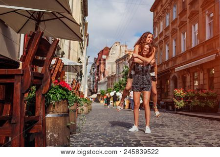 Young Woman Carrying Her Best Friend On Her Back On City Street. Happy Girls Students Laughing And H