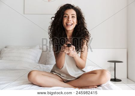 Photo of attractive caucasian woman with curly hair looking at camera and holding remote control while sitting in bed at home on white bedding with legs crossed