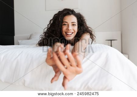 Portrait of prettyy european woman with long curly hair lying in bed with white linen in apartment and stretching arms to you with smile