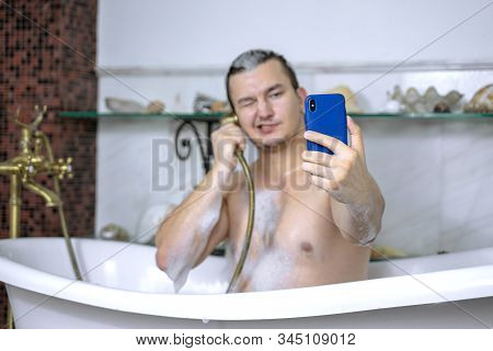 Young Handsome Naked Caucasian Man Blogger In Bathtub Makes Selfie On Mobile Phone, With Bronze Fauc