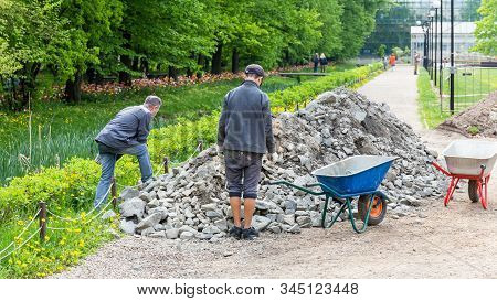 Two Workers Load Stones Into A Wheelbarrow During The Repair Of The Sidewalk In The Park