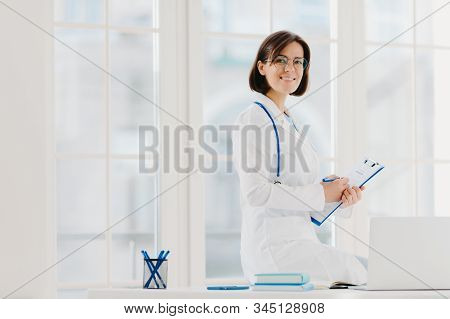 Skilled Female Physician Writes In Clipboard, Poses At Workplace With Laptop Computer. Professional 