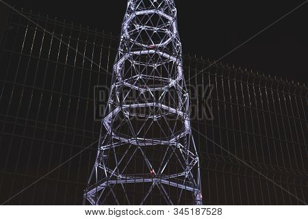 Night View Of The Illuminated Base Of A Television Or Radio Tower Located Behind A Trellised Fence A