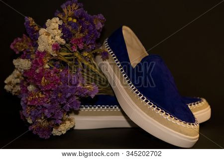 On Black Background, Pair Of Shoes, Blue Suede Slipons With White Thick Sole. Bouquet Of Dry Field F