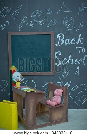 Happy Smiling Pupil Drawing At The Desk. Class Room With Blackboard On Background. Alarm Clock, Penc